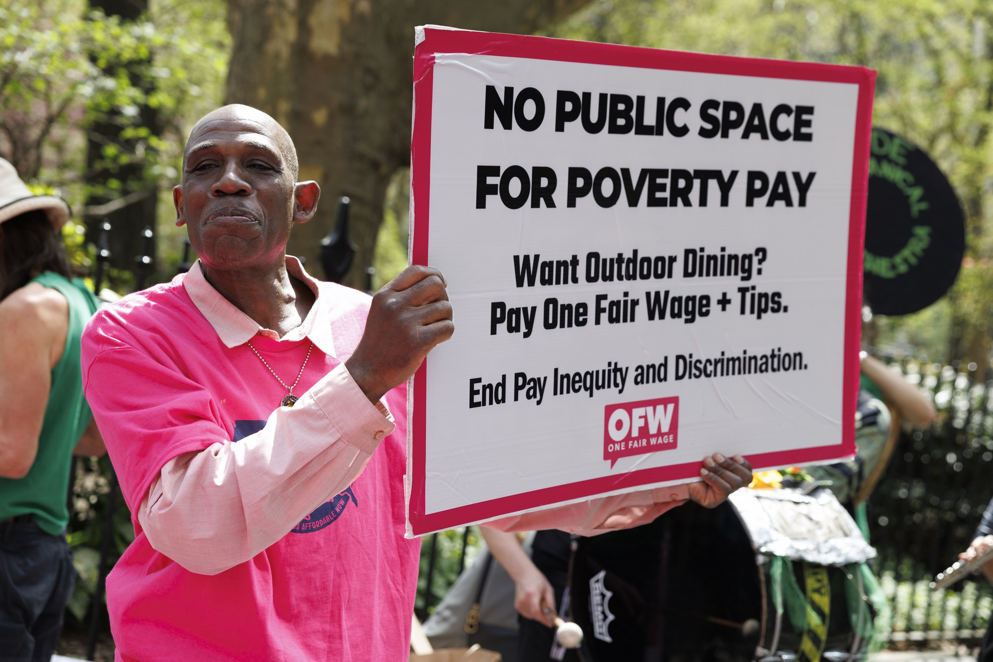 An OFW member holds a placard reading &lsquo;NO PUBLIC SPACE FOR POVERTY PAY &mdash; Want Outdoor Dining? Pay One Fair Wage + Tips. End Pay Inequity and Discrimination.&rsquo; at the OFW action at NYC City Hall, April 16, 2026